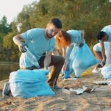 Voluntari cu mănuși și saci care strâng deșeuri de pe malul unui canal mărginit de stuf